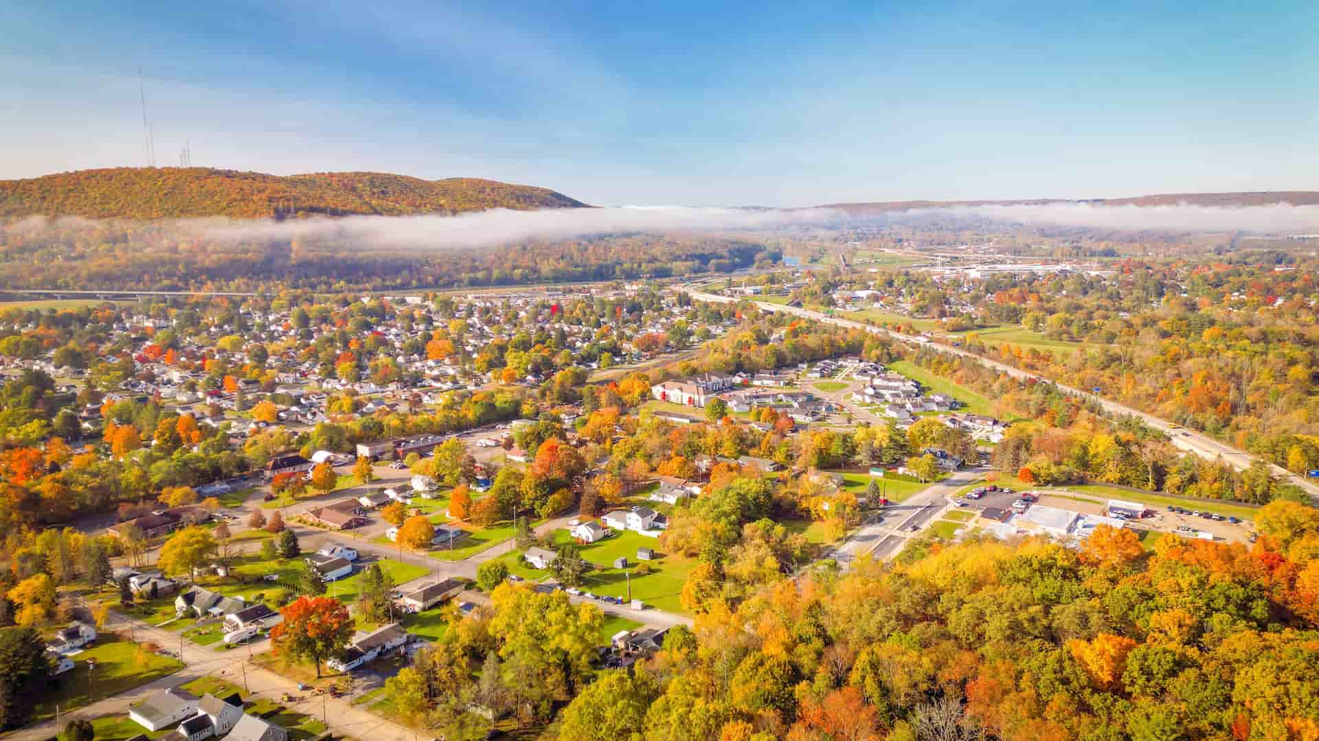 "An aerial view of the town of Corning, New York, in the autumn, with houses and streets surrounded by a vibrant tapestry of orange and yellow foliage, and low-lying clouds hanging over the forested hills in the distance. "