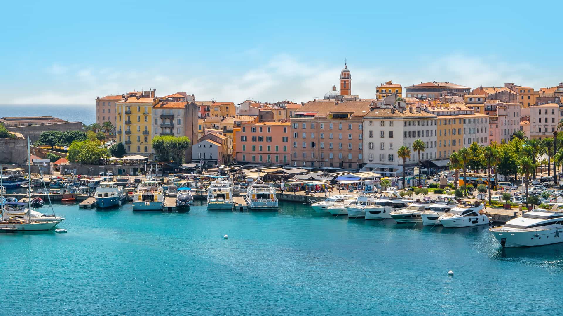 A beautiful view of the bustling harbor in Ajaccio, Corsica, with colorful historic buildings lining the port and numerous yachts docked in the marina.