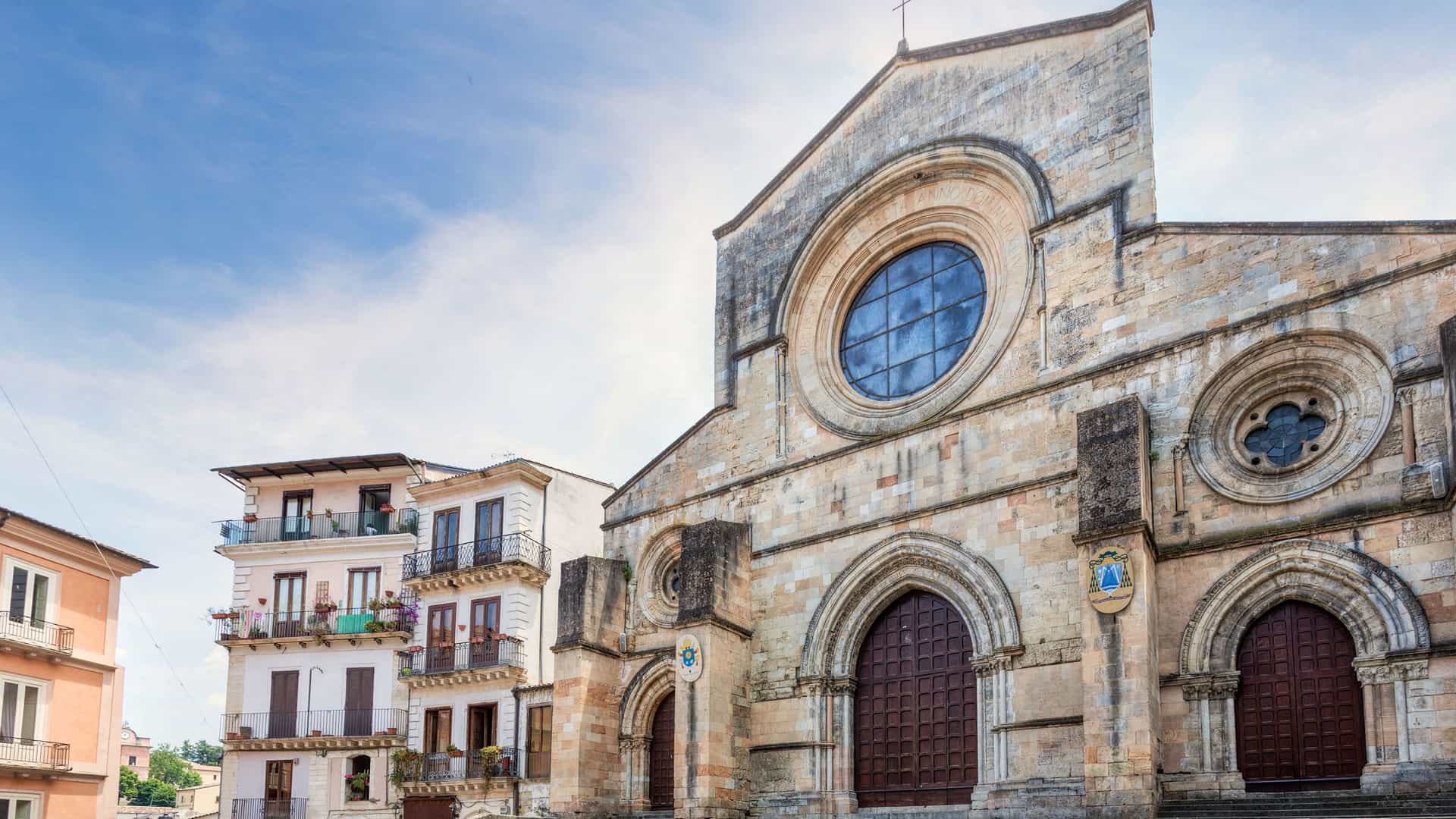 A historic cathedral in Crotone, Italy, with two large arched doorways, an ornate circular window, and a modern apartment building to its left.