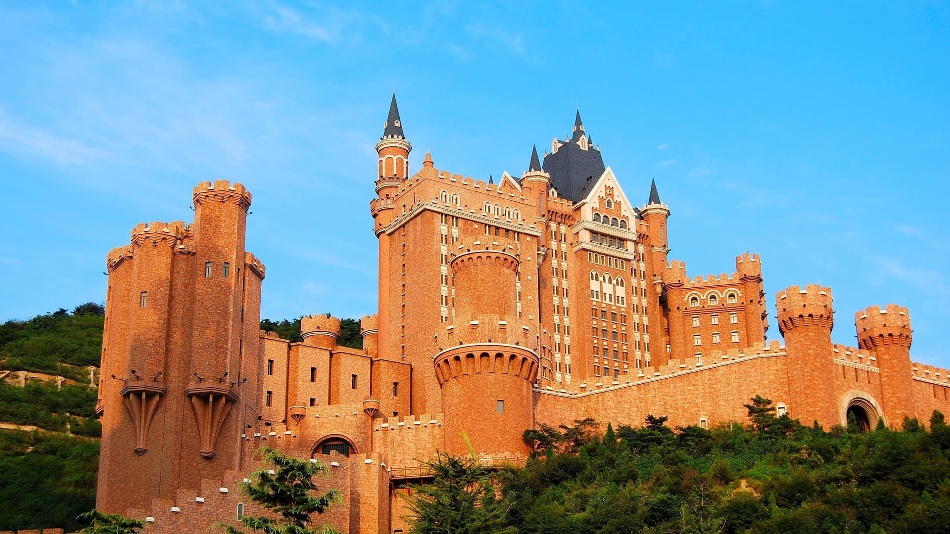 A majestic modern castle, the Dalian Castle Hotel, with its red brick facade and multiple turrets, stands on a hillside in Dalian, China.
