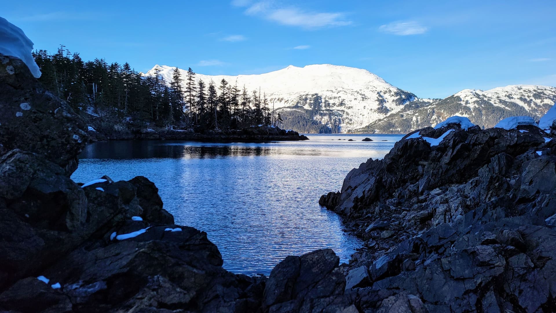 A scenic view of the calm waters of Decision Passage, with a snow-dusted, forested shoreline and majestic snow-capped mountains in the background.