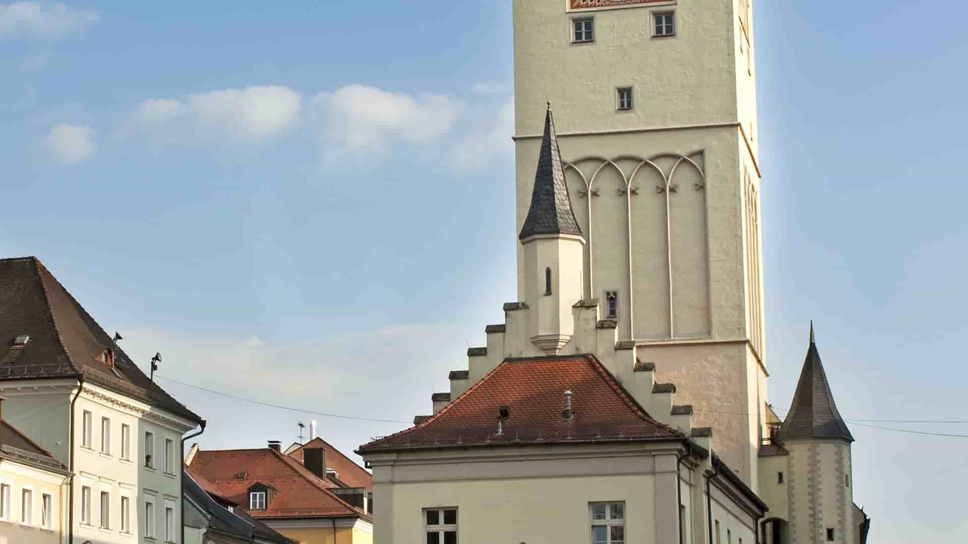 The historic Rathaus tower in Deggendorf, Germany, with its distinctive stepped gable and pointed roof, against a bright blue sky.
