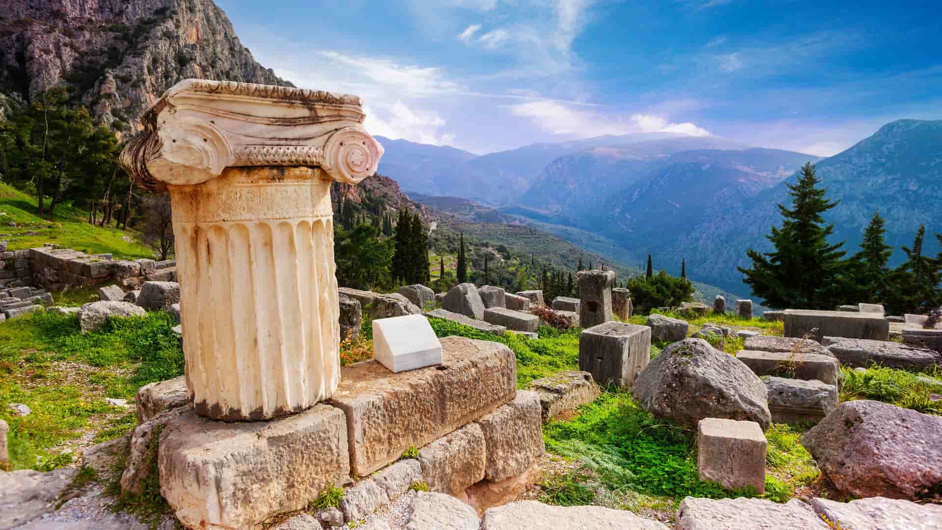 The ancient Greek ruins of Delphi, Greece, featuring a large column fragment in the foreground with a dramatic mountain and valley backdrop.