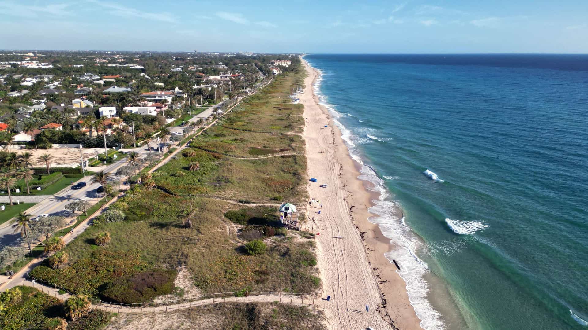 "An aerial view of the beautiful sandy beach and turquoise ocean of Delray Beach, Florida, with a lifeguard stand in the middle ground and a line of palm trees and coastal homes on the shoreline. "