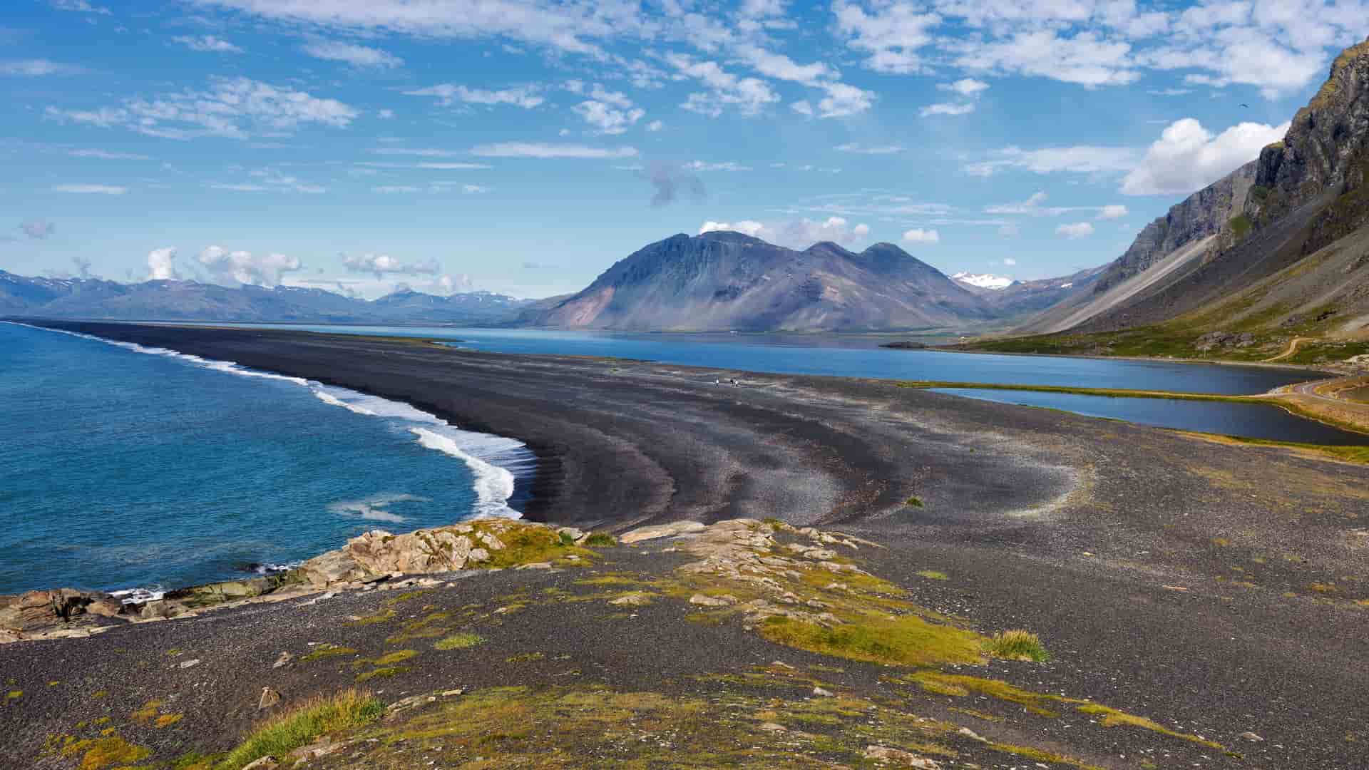 A panoramic view of the dramatic black sand beach and rugged coastline near Djúpivogur, Iceland, with a sweeping mountain range in the background.