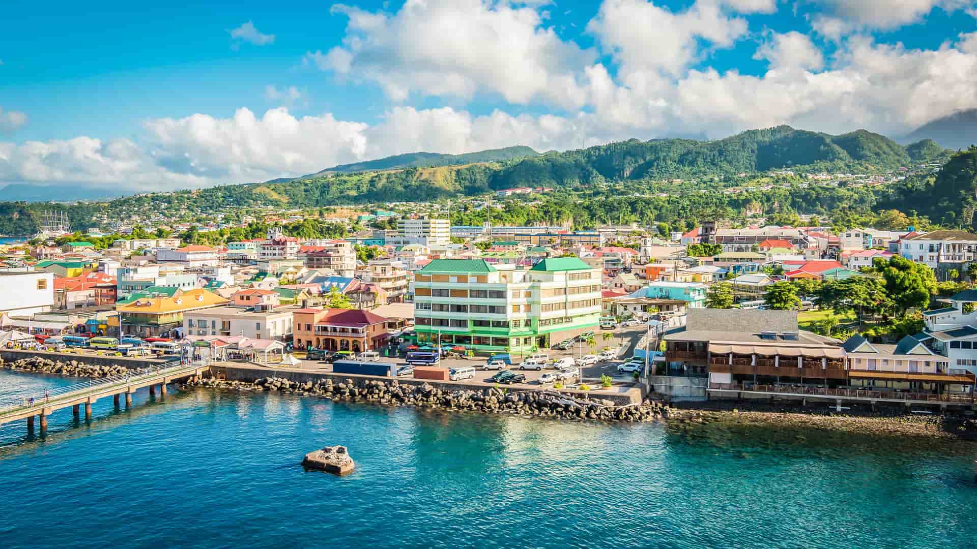 A panoramic view of the coastal city of Roseau in Dominica, with its colorful buildings nestled between the Caribbean Sea and lush, mountainous terrain under a beautiful sky.