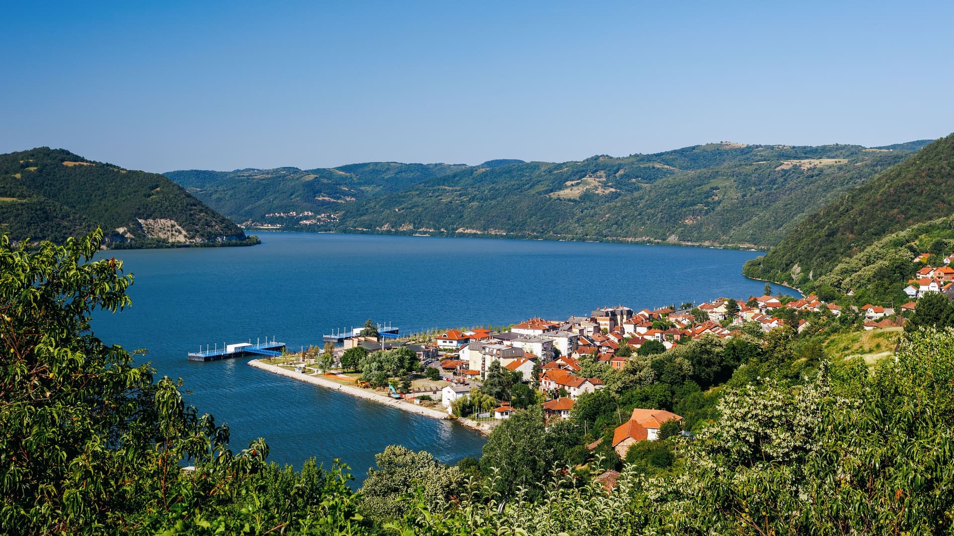 The picturesque town of Donji Milanovac, Serbia, nestled on the wide banks of the Danube River, surrounded by the lush, green hills of Đerdap National Park.