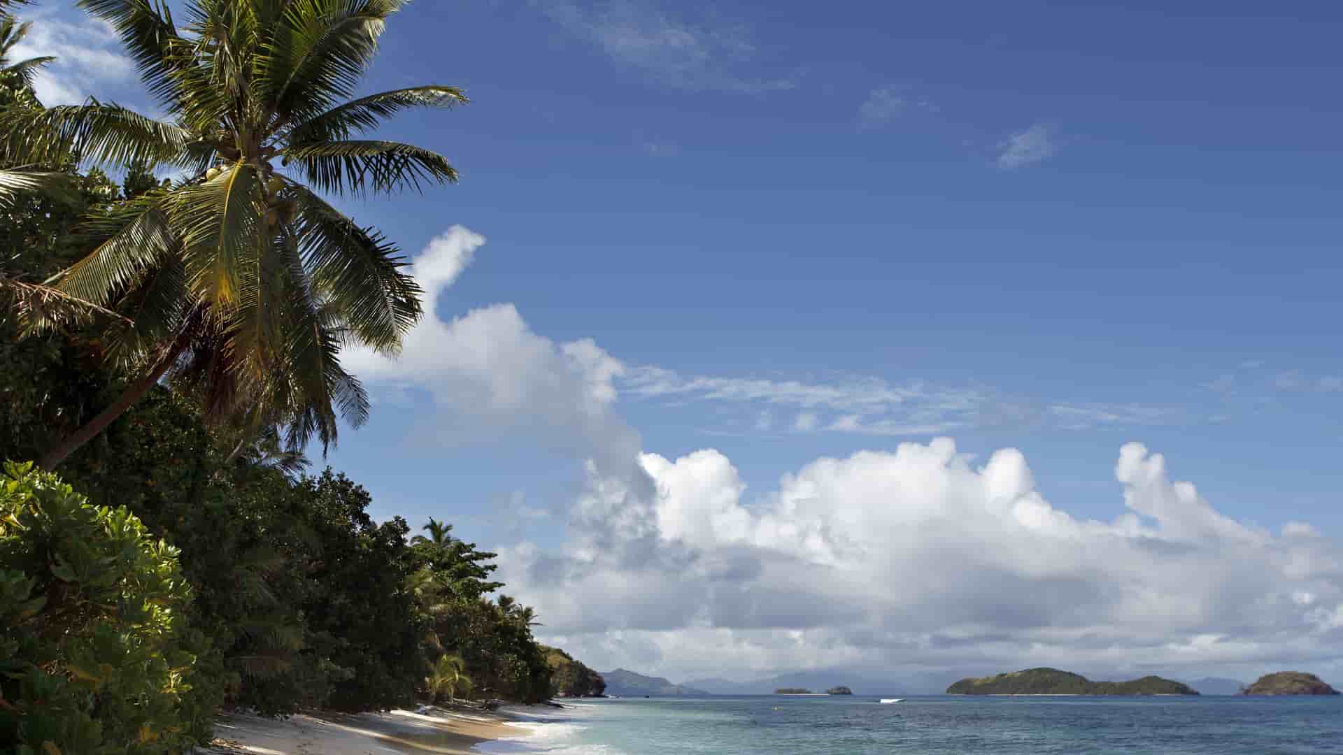 A scenic tropical beach on Dravuni Island, Fiji, with a pristine white sand shoreline, turquoise water, lush palm trees, and small islands in the distance.