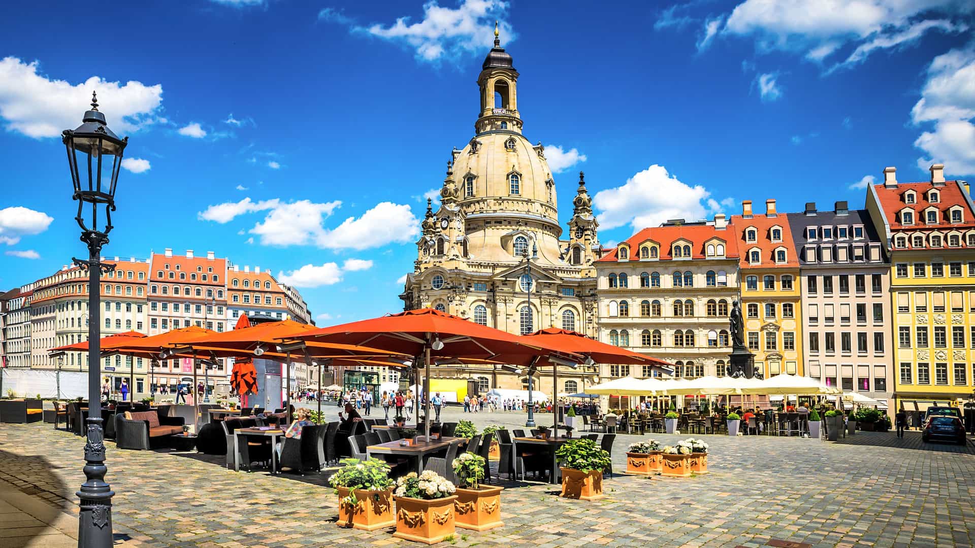 A sunny view of a cafe with orange umbrellas in the square of Dresden, Germany, with the historic Frauenkirche and other baroque buildings in the background.