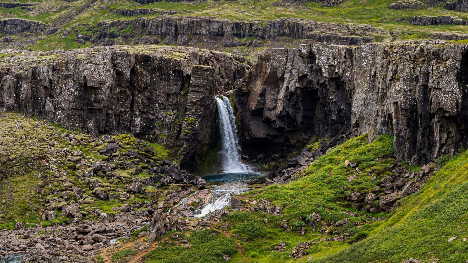 "A powerful waterfall cascading over a cliff face into a small turquoise pool, surrounded by rugged, rocky cliffs and vibrant green moss in the scenic landscape near Egilsstaðir, Iceland. "