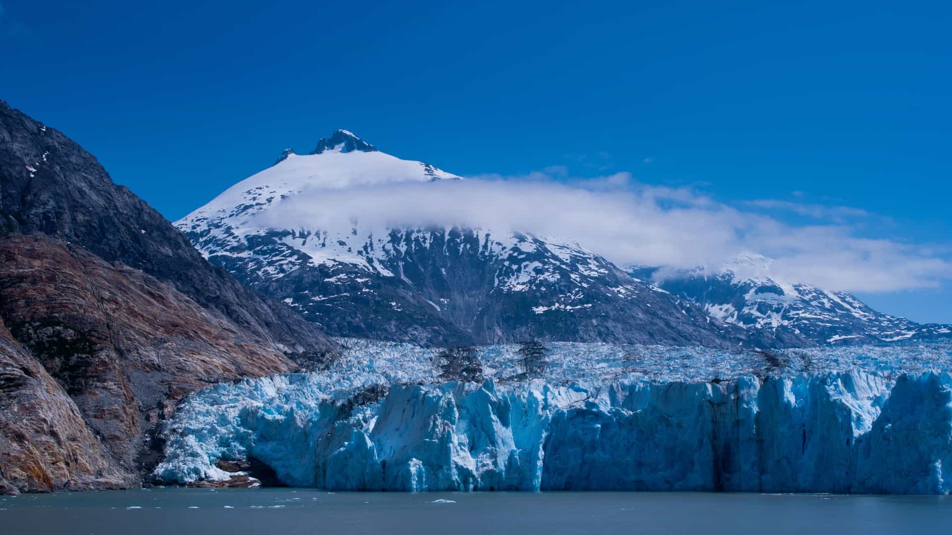 A dramatic view of a towering blue glacier and a jagged, snow-capped mountain at Endicott Arm, Alaska, with clear blue water and sky in the stunning landscape.