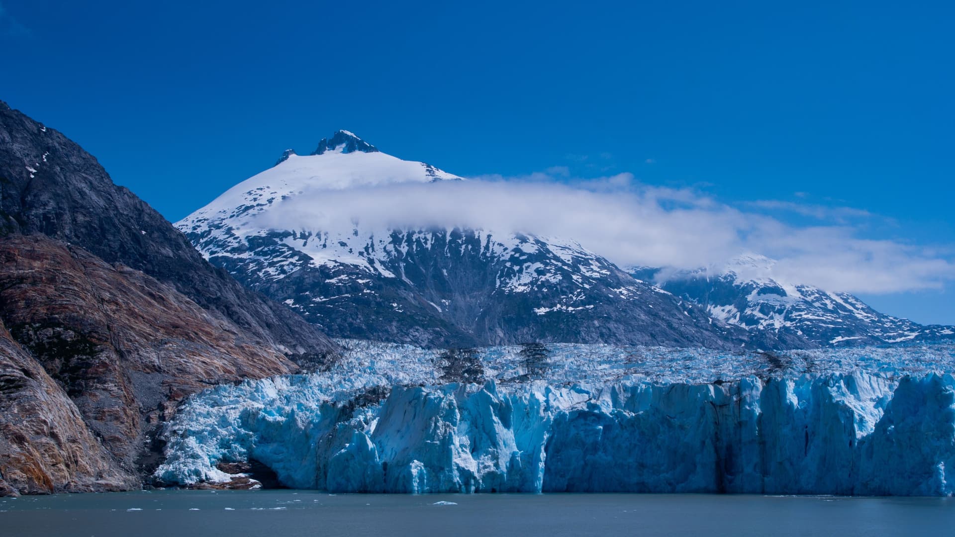 A dramatic view of a towering blue glacier and a jagged, snow-capped mountain at Endicott Arm, Alaska, with clear blue water and sky in the stunning landscape.