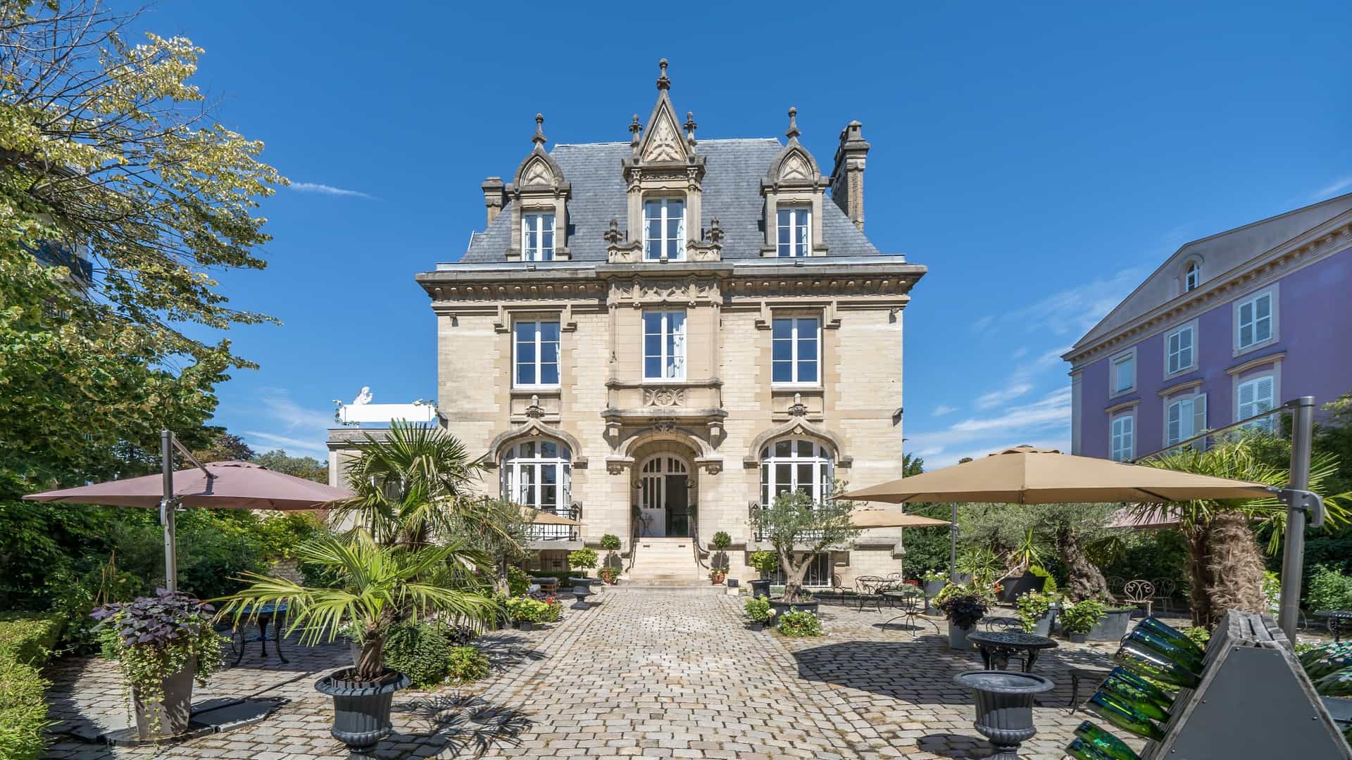 A large, ornate historic manor house with a grey slate roof in Epernay, France, with a well-manicured cobblestone courtyard, manicured plants, and outdoor seating.