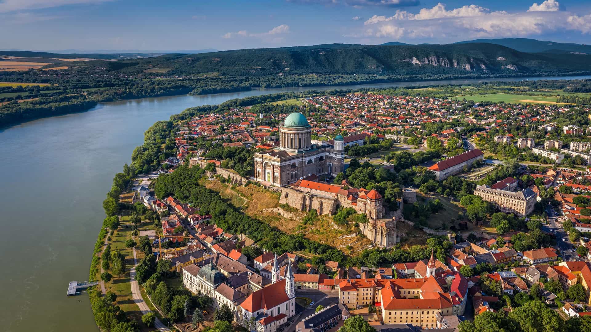 An aerial view of Esztergom Basilica and Esztergom Castle on a hilltop overlooking the Danube River and the town below, with lush green hills in the background.