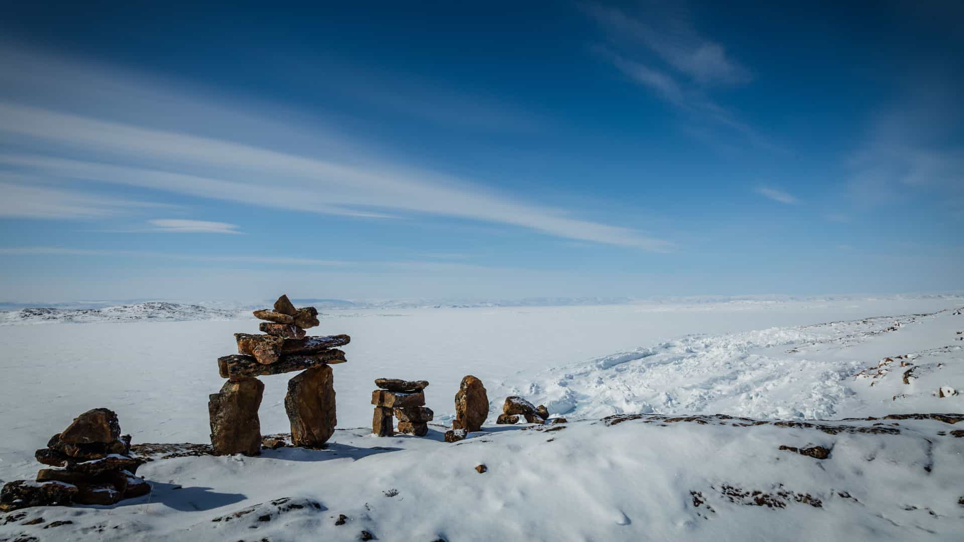 A snowy Arctic landscape in Flexure Bay featuring several stacked stone inuksuit standing on a wide expanse of snow, with distant hills beneath a bright blue sky scattered with soft clouds.