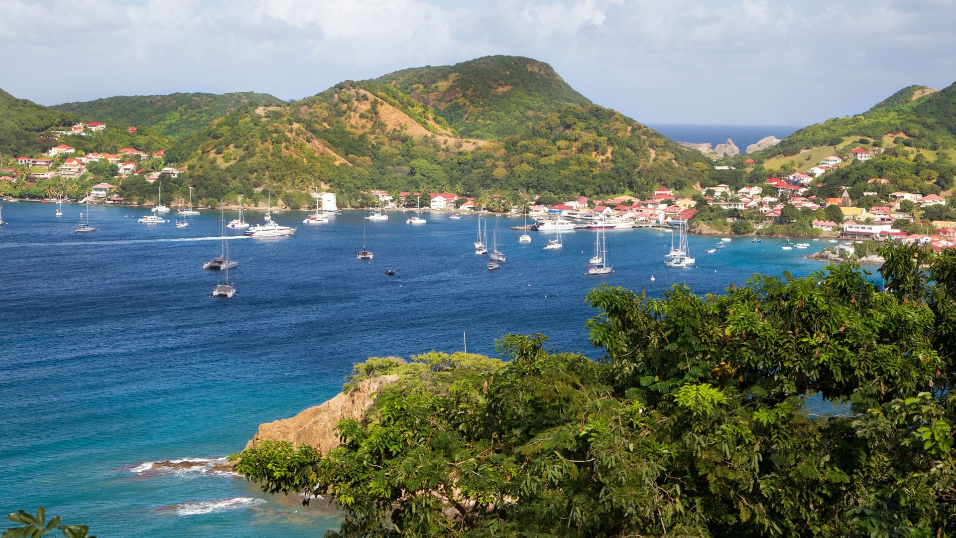 A panoramic view of Fort-de-France, Martinique, showing a bustling bay filled with boats and yachts, with colorful houses nestled on the surrounding green hillsides and the deep blue Caribbean Sea beyond.