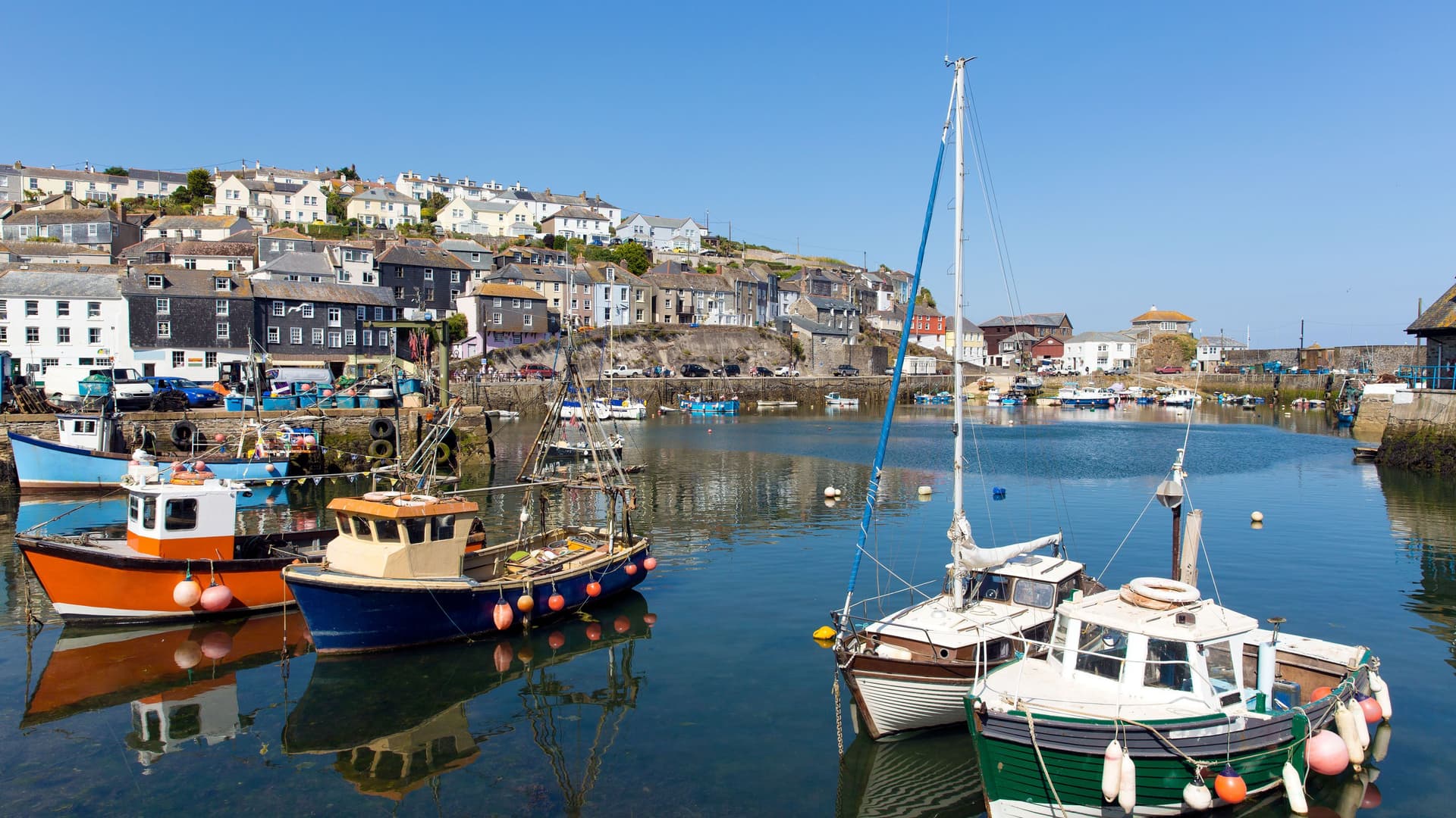 A bright, sunlit harbor scene in Fowey, Cornwall, featuring colorful fishing boats and yachts moored in the calm water with the town's buildings rising up the hill in the background.