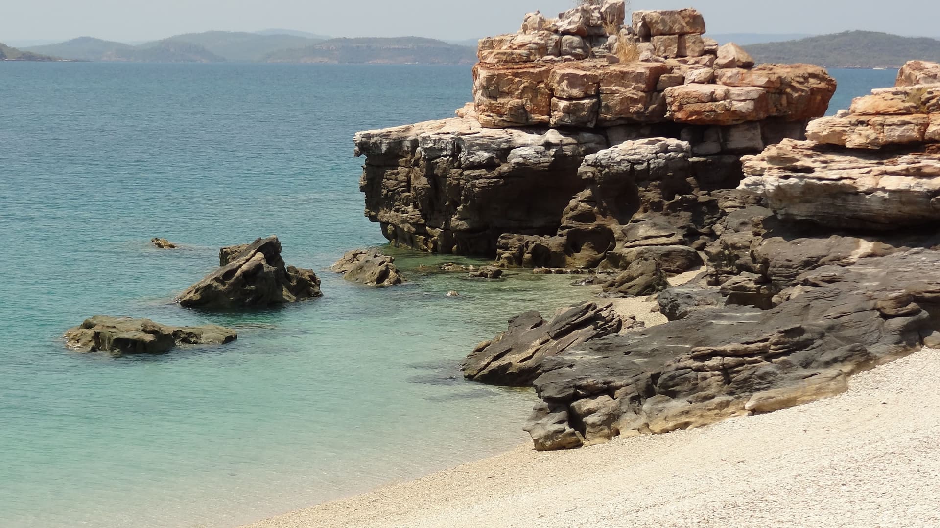 A close-up view of the rugged, layered rock formations and sandy shore of Freshwater Cove, Australia, with a tranquil turquoise ocean and distant hills under a clear sky.
