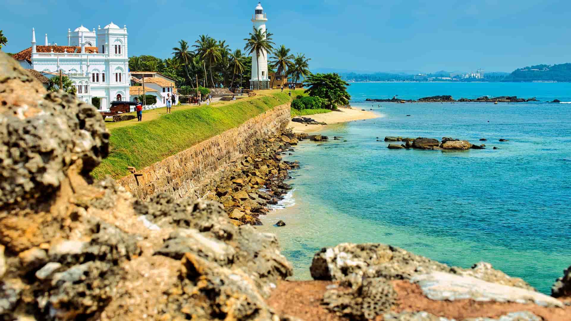 A beautiful panoramic view of the Galle Fort in Sri Lanka, featuring a historic white lighthouse and buildings perched on a rocky coastline overlooking the tranquil Indian Ocean.