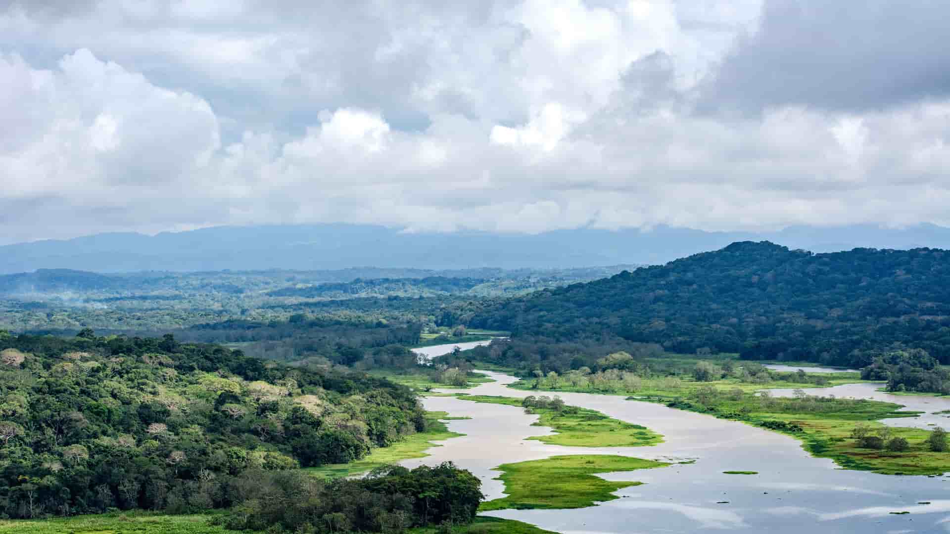 "A scenic aerial shot of the Chagres River winding through the dense, green rainforest of Gamboa, Panama, with mountains in the misty distance and a cloudy sky above. "