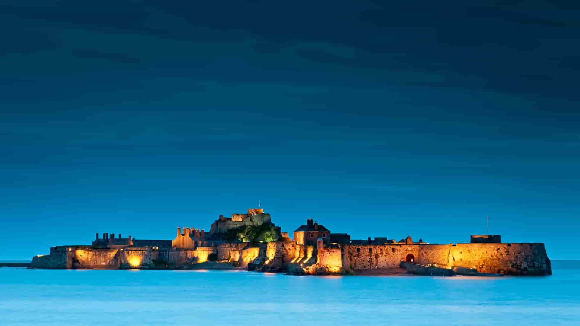 A historic castle fortress illuminated at night, located on a small island surrounded by calm water under a deep blue evening sky.