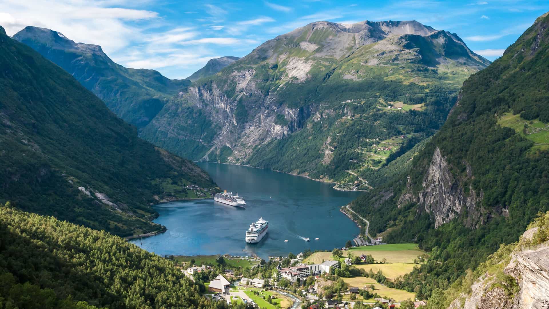 A panoramic view of the majestic Geirangerfjord in Norway, showcasing two large cruise ships docked in the calm waters of the fjord surrounded by towering, green mountains.