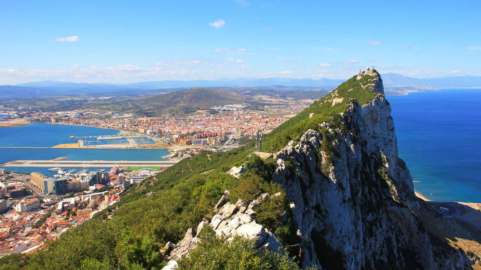 A scenic panoramic shot from the top of the Rock of Gibraltar, overlooking the city, marina, and the Mediterranean Sea under a bright blue sky.