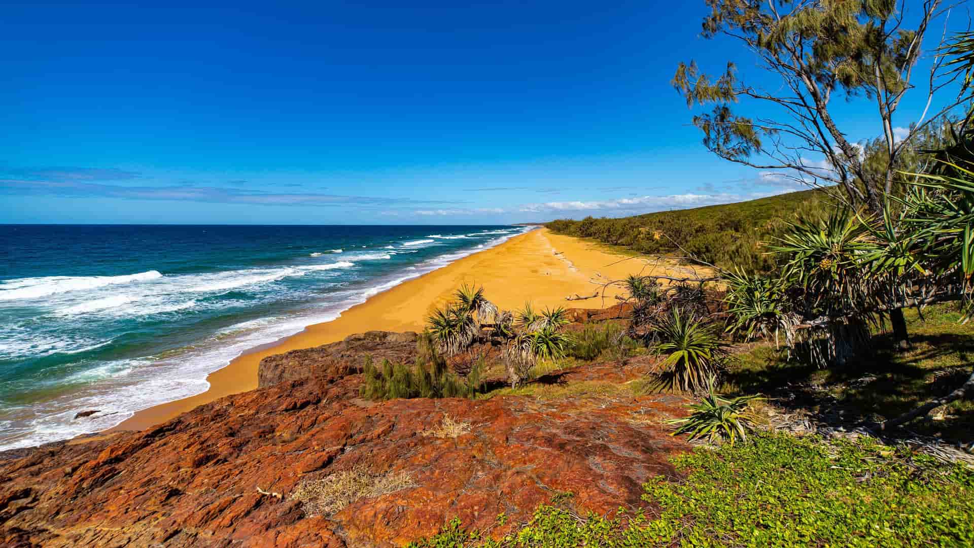 A scenic shot of the Gladstone coastline in Queensland, Australia, showcasing a pristine sandy beach, red rocky cliffs, and a beautiful blue ocean under a clear sky.