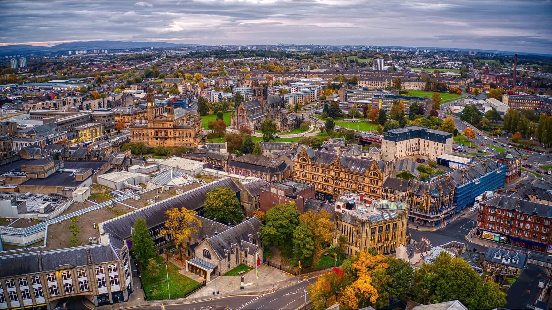 An aerial shot of Glasgow, Scotland, featuring the city's historic architecture, including the University of Glasgow, and a large urban landscape filled with autumn-colored trees under a dramatic, cloudy sky.