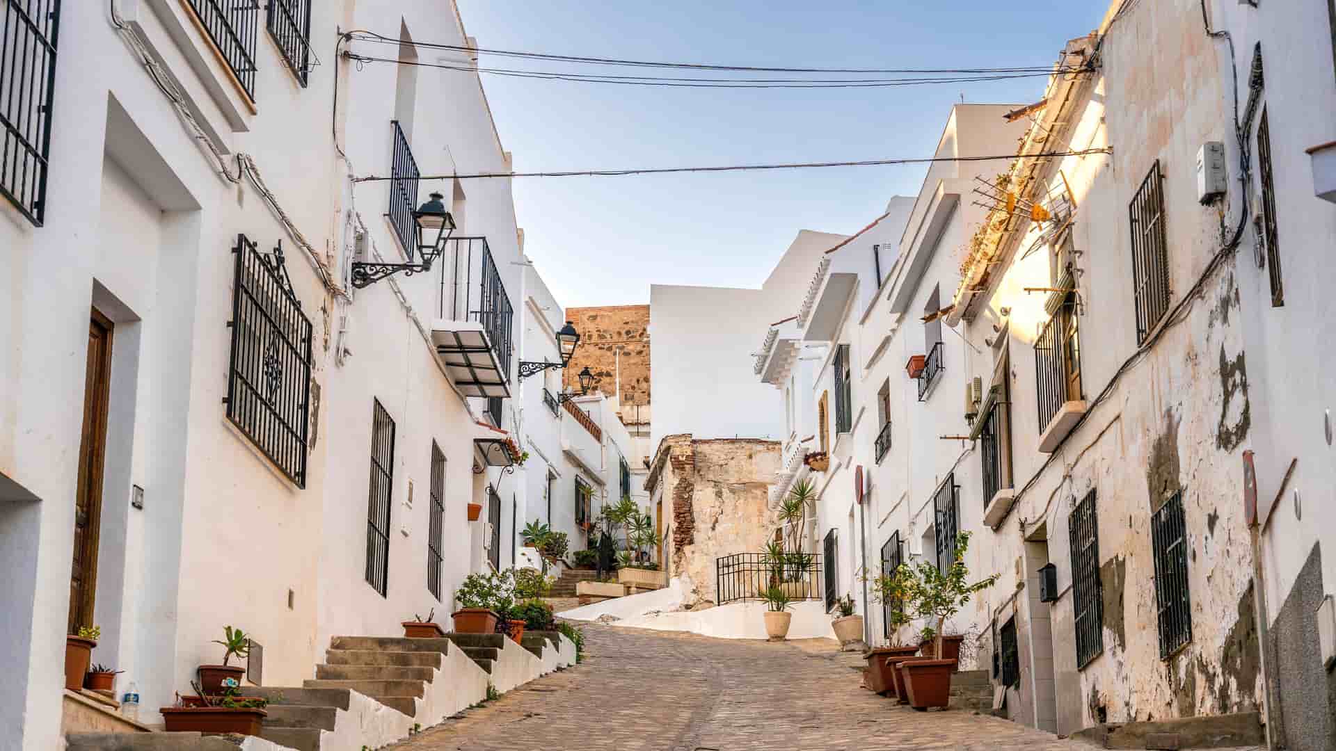 A picturesque view of a narrow, cobblestone street in a historic white-washed village near Motril, Spain, lined with buildings and potted plants.