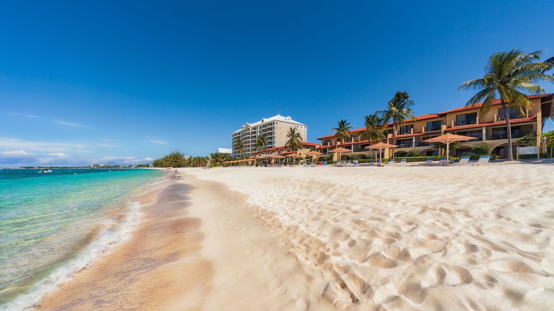 A scenic shot of the stunning Seven Mile Beach in Grand Cayman, with a wide, sandy beach, calm turquoise water, and resorts with palm trees under a beautiful blue sky.