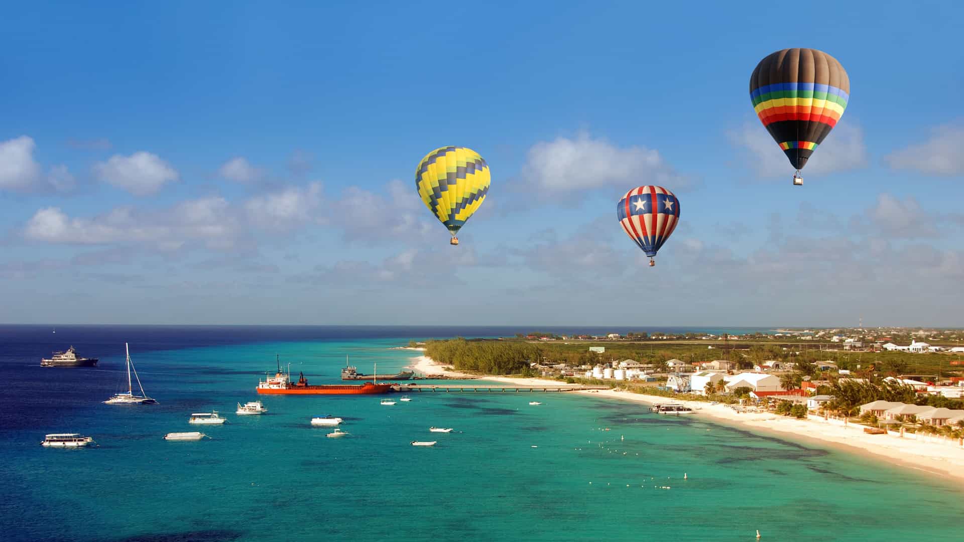 A sunny, tropical beach on Grand Turk Island with multiple hot air balloons flying over the turquoise water and shoreline.