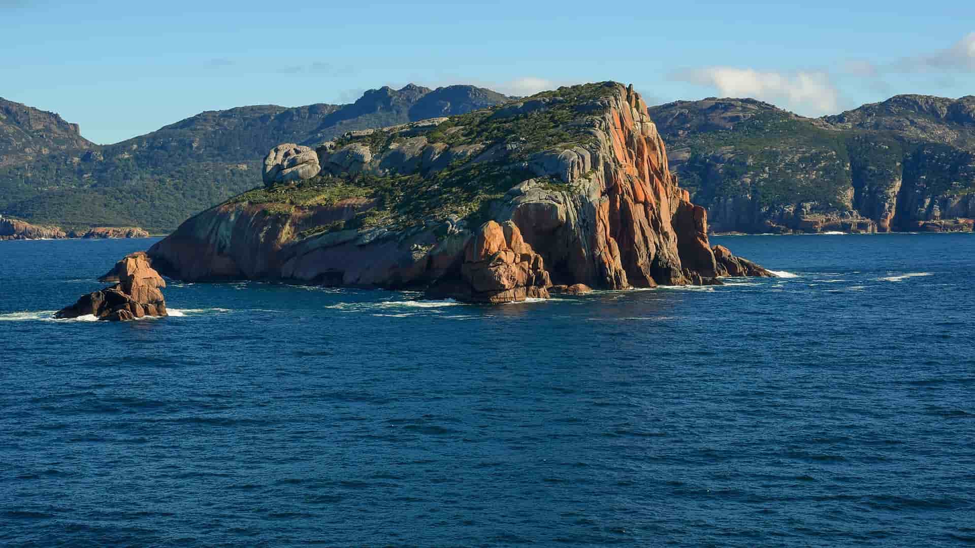 A wide shot of the dramatic, rocky coastline of Freycinet National Park, with a large, distinctively orange-red granite sea stack jutting out of the calm, blue waters of Great Oyster Bay in Tasmania, Australia.