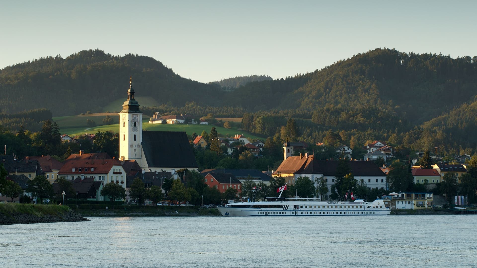 A river cruise ship docked in the picturesque town of Grein, Austria, with a prominent church steeple and residential buildings nestled against lush green hills.