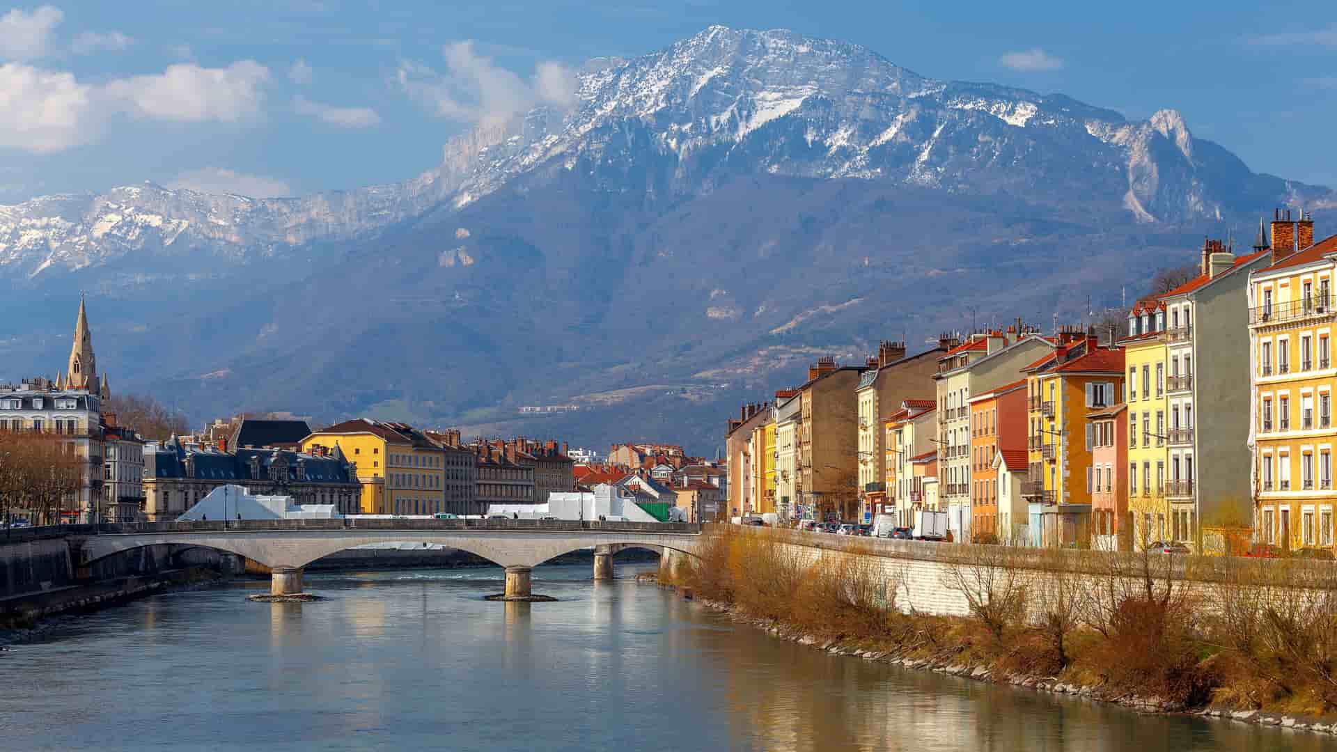 "A scenic view of Grenoble, France, with the Isère River flowing through the city, a bridge connecting the two sides, a row of colorful buildings, and a backdrop of the majestic, snow-dusted French Alps. "