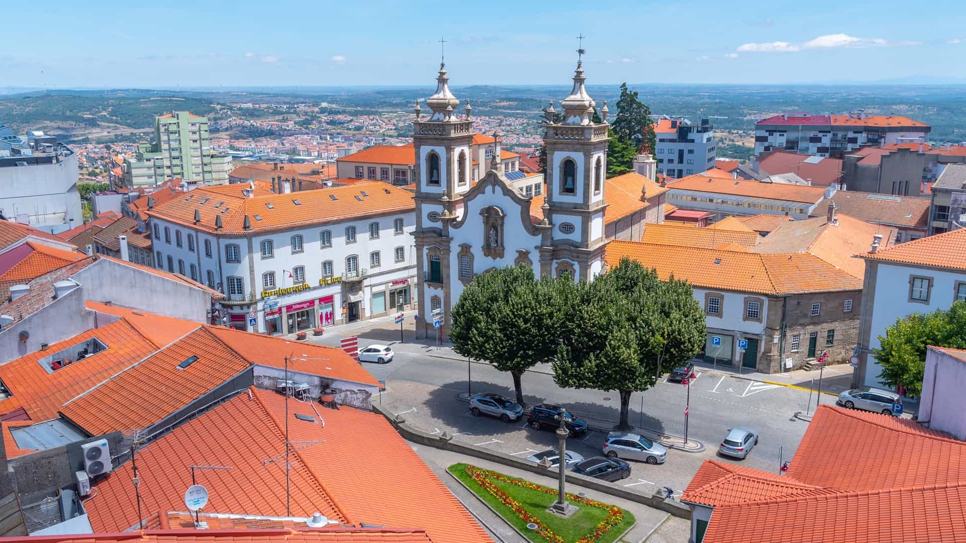 "An aerial view of the historic city of Guarda, Portugal, with a church featuring two ornate bell towers at its center, surrounded by a mix of red-roofed buildings and modern architecture. "