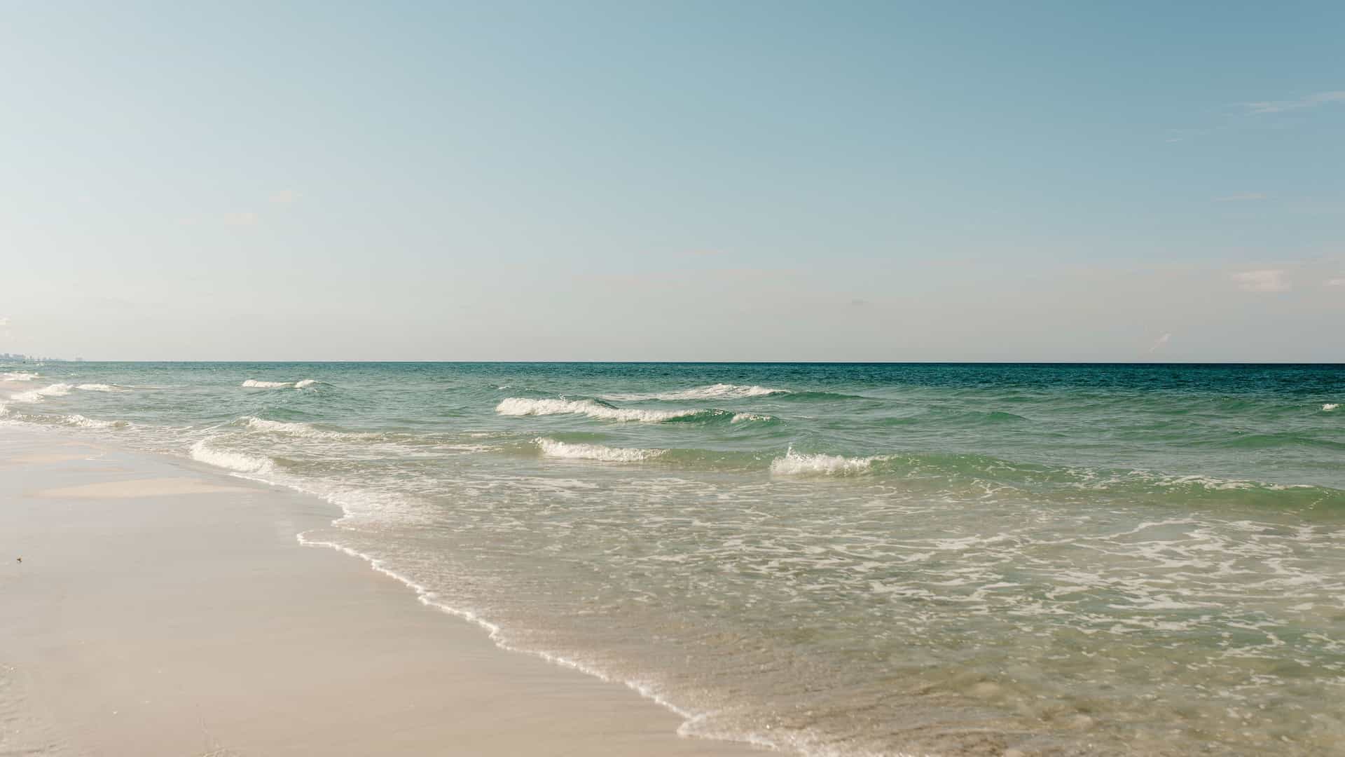 A tranquil wide-angle shot of a sandy beach along the Gulf of Mexico, with gentle waves rolling onto the shore and a clear blue sky.