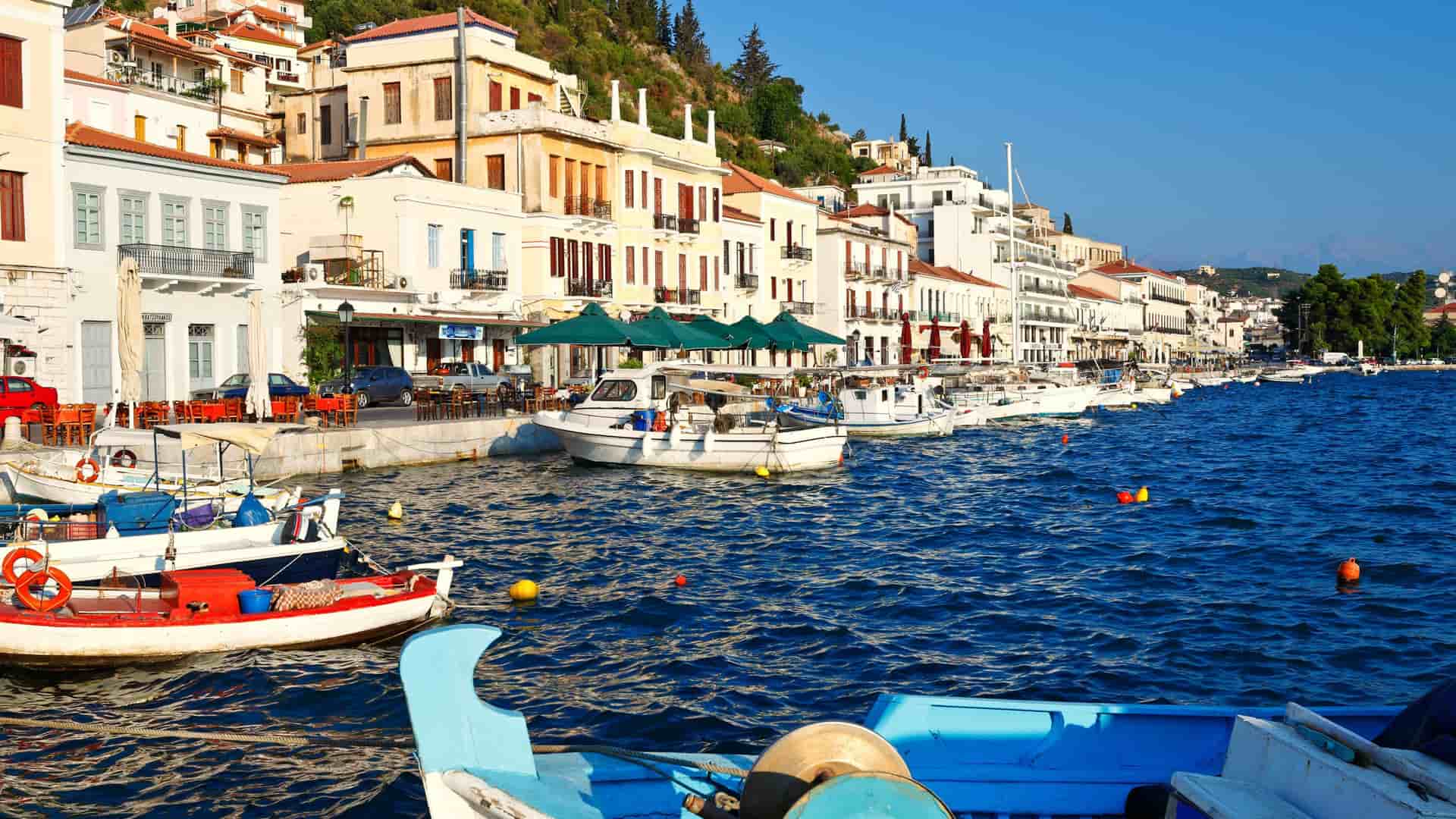 A scenic view of the waterfront in Gythion, Greece, with colorful fishing boats and yachts docked in the harbor next to traditional white buildings with orange-tiled roofs.