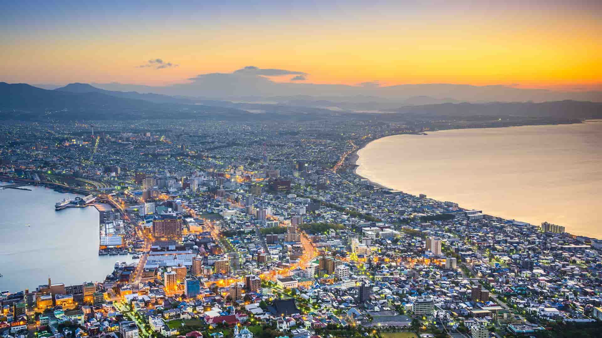 A stunning high-angle, aerial shot of Hakodate, Japan, at dusk, with city lights illuminating the urban sprawl nestled between two bays and a mountain range in the distance.