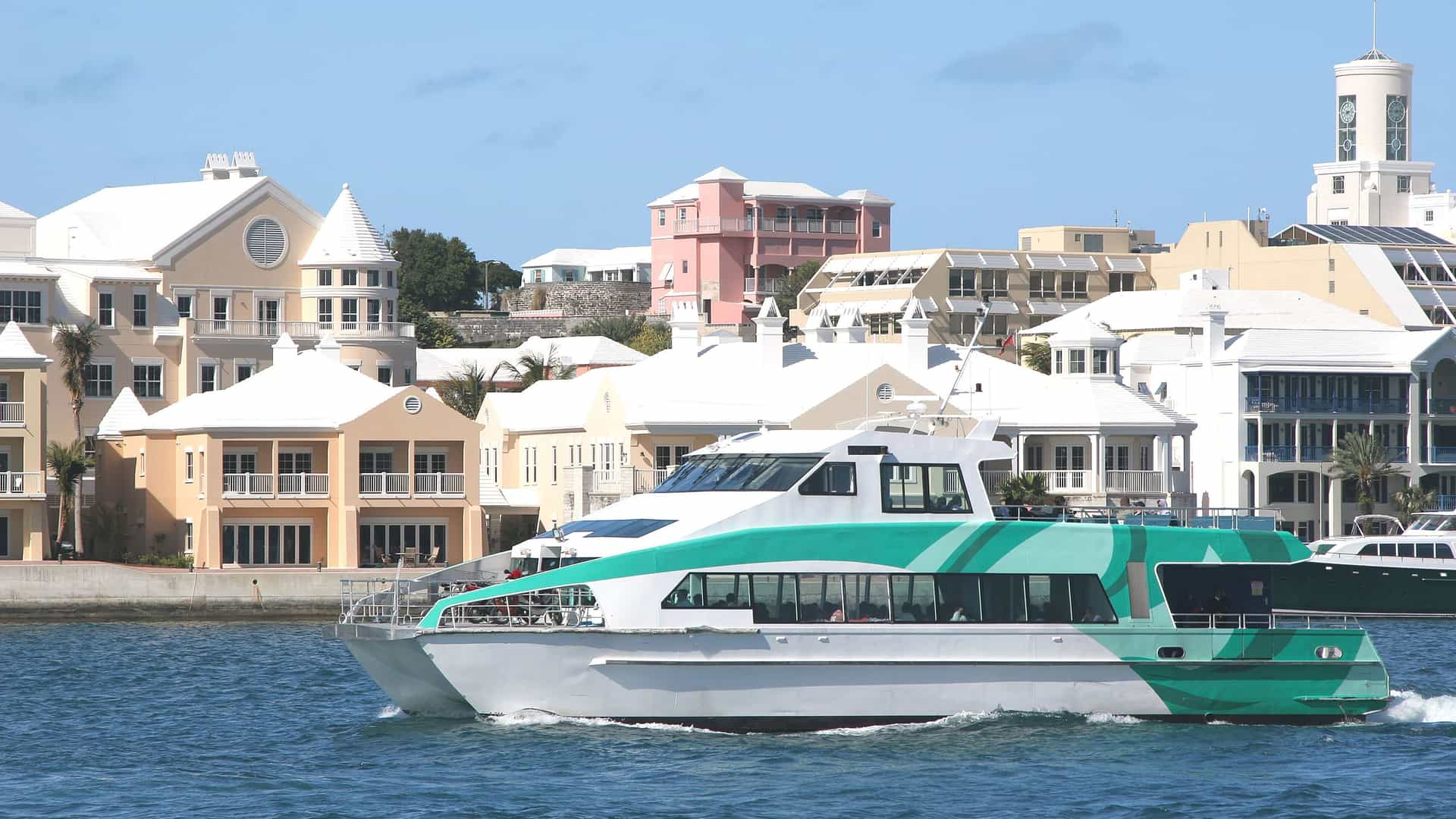A turquoise and white ferry boat cruises through the harbor in Hamilton, Bermuda, with a backdrop of pastel-colored buildings and houses along the waterfront.