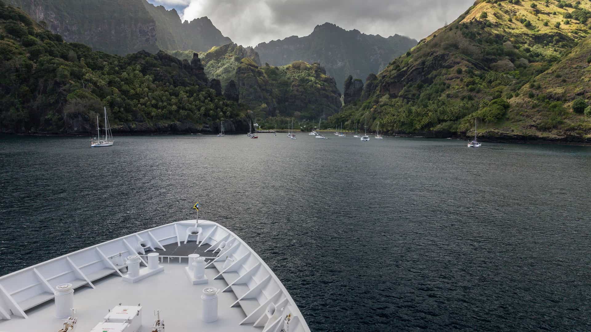 A dramatic view from the bow of a ship entering Hanavave Bay, Fatu Hiva, with numerous sailboats anchored in the dark water surrounded by lush, steep volcanic mountains.