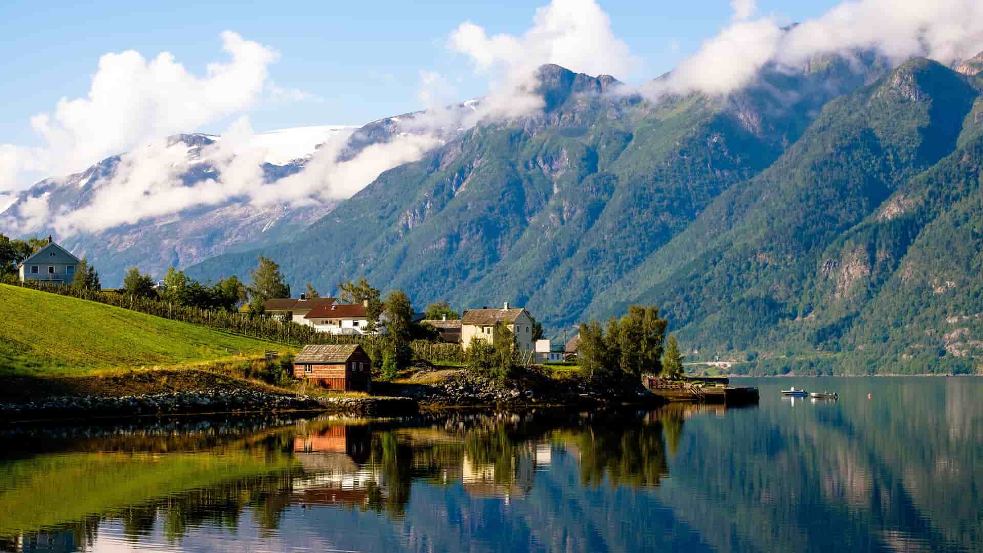 A picturesque view of Hardangerfjord, Norway, with small houses on a lush, green hillside reflected in the calm water and towering snow-capped mountains in the background.