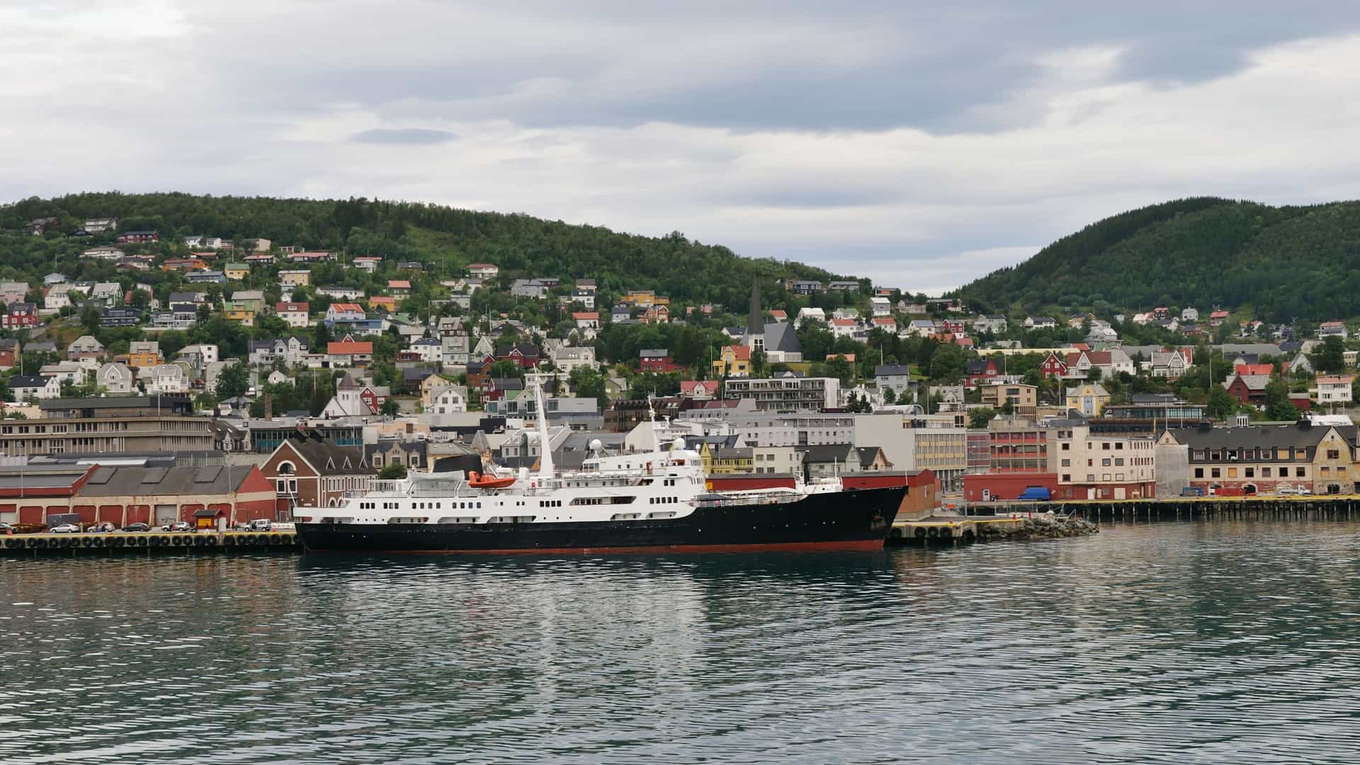 A large black and white ship is docked at the harbor in Harstad, Norway, with the city's colorful houses sprawling up the lush green hillsides in the background.