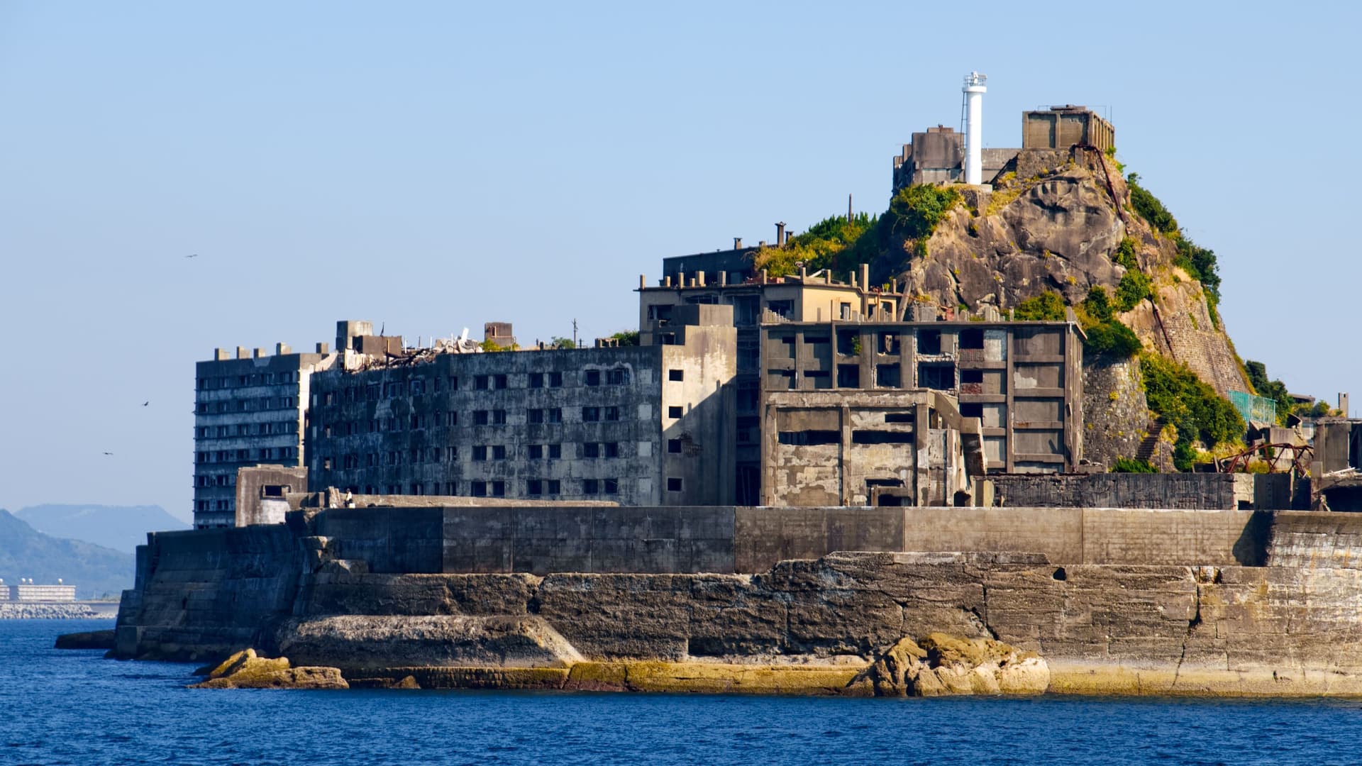 A haunting view of Hashima Island, also known as Gunkanjima, a deserted city with derelict concrete buildings and a lighthouse standing on a rocky outcrop in the sea.