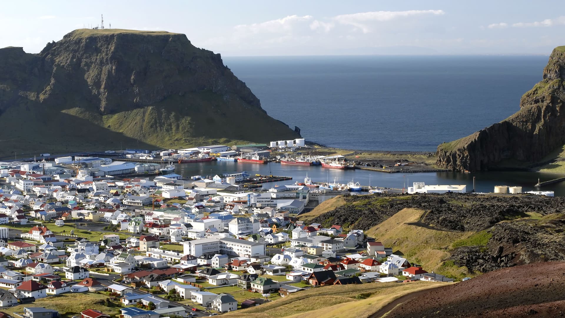 A scenic view of the town of Heimaey, Iceland, with a harbor filled with fishing boats, colorful houses, and two large volcanic rock formations surrounding the bay.