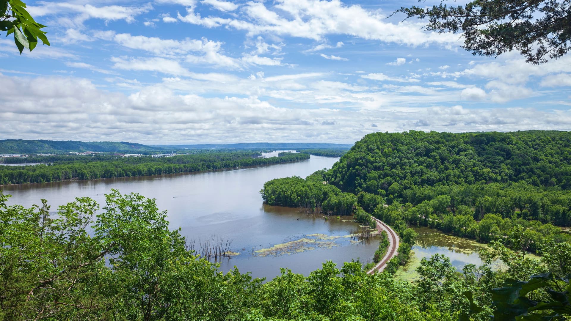 A panoramic, high-angle view of a wide river with a forested island in the middle, and a winding road leading to it. The sky is bright blue with scattered clouds.
