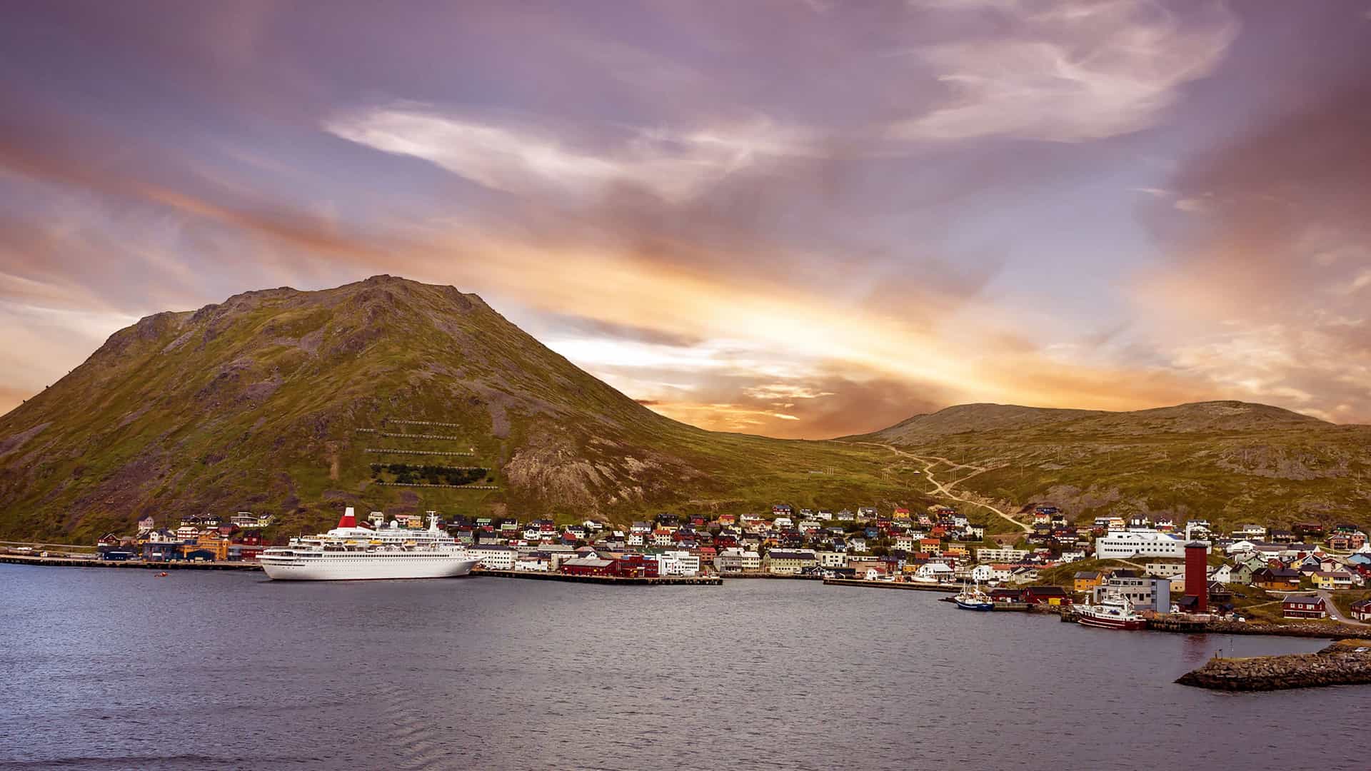 A cruise ship docked in the port of Honningsvag, Norway, with the colorful harbor town nestled at the base of a large, grassy mountain under a dramatic sky.