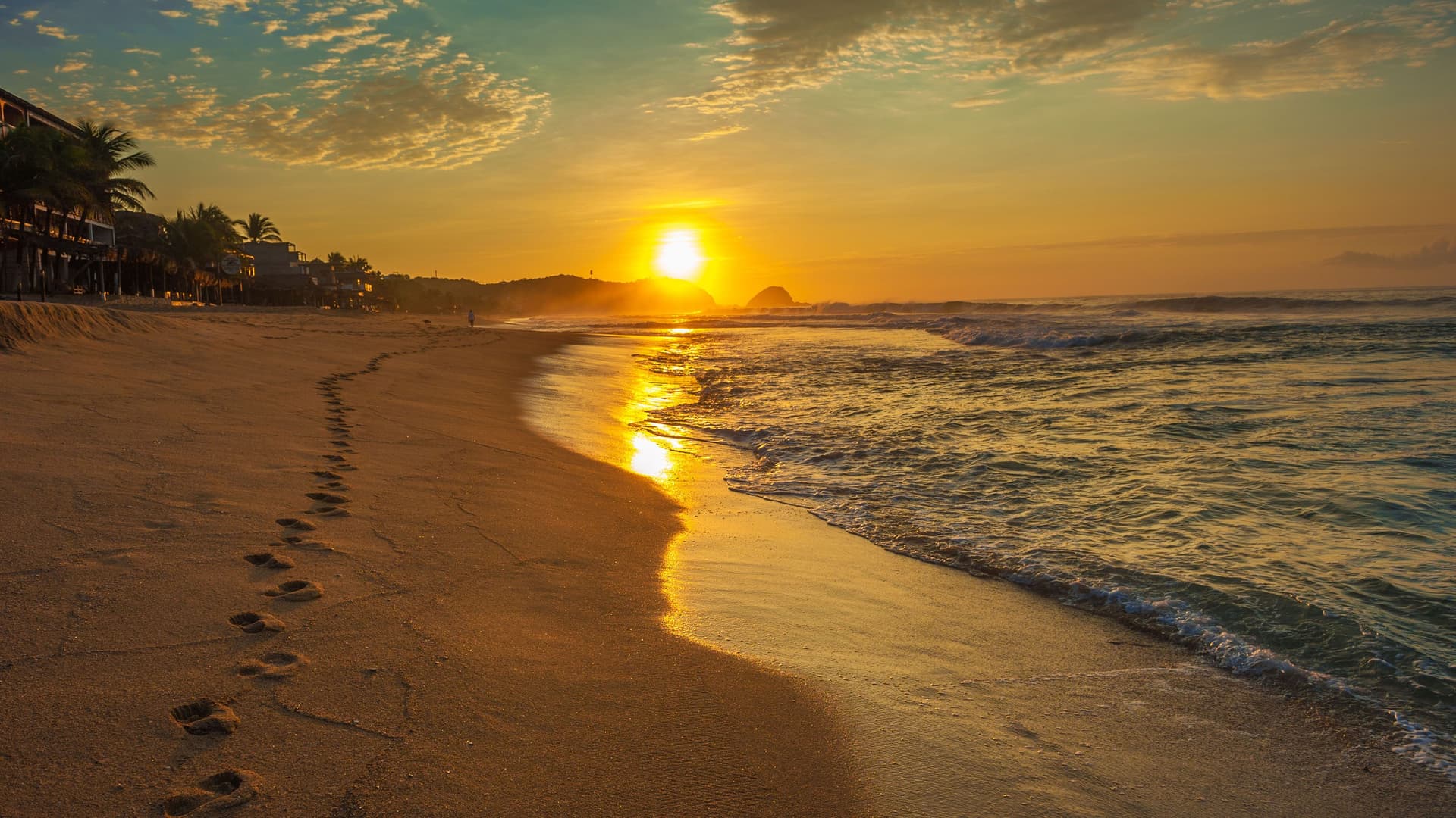A golden sunset over the beach in Huatulco, Mexico, with footprints in the sand leading towards the shimmering ocean and a warm glow over the water.