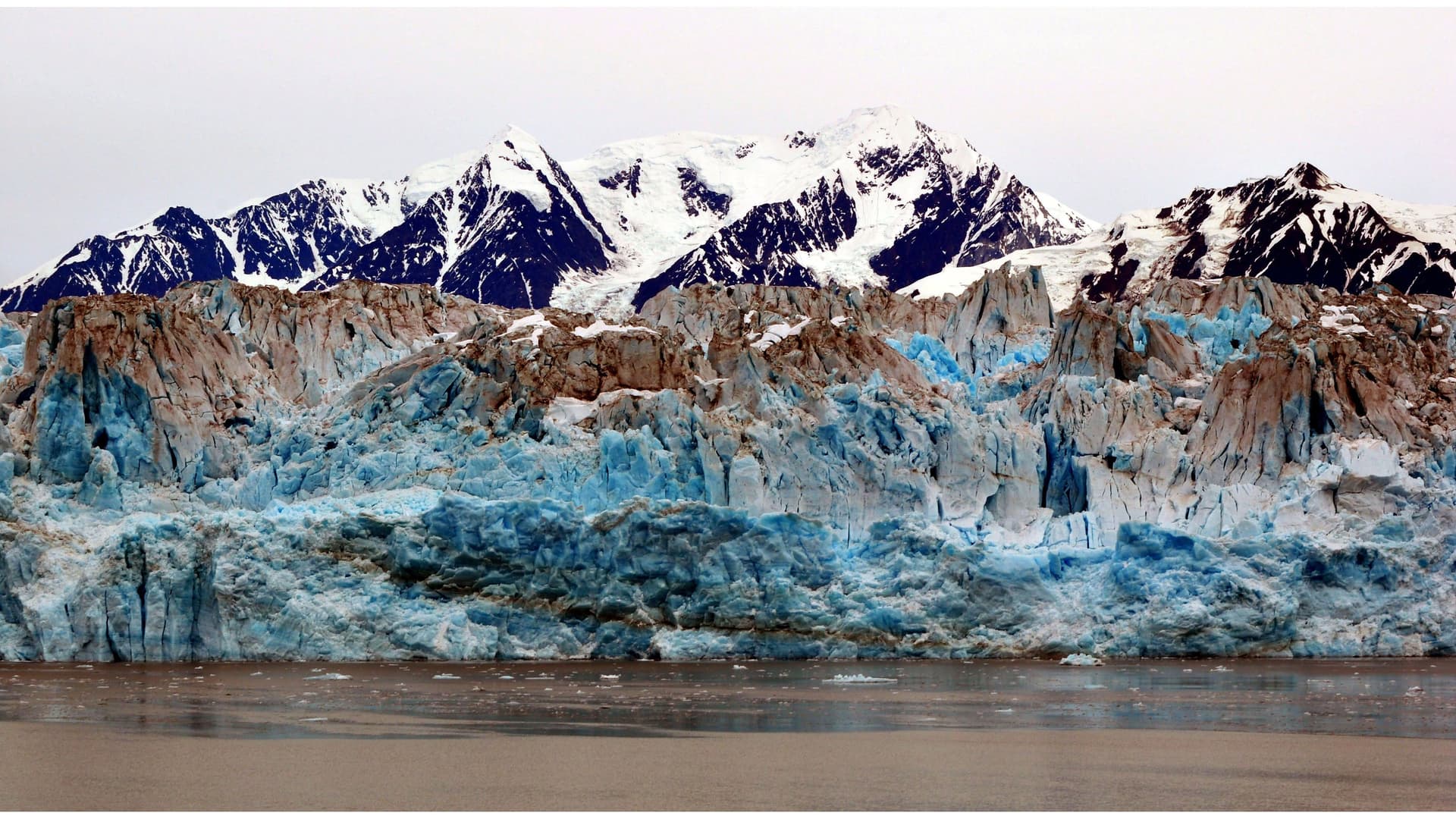 A stunning close-up of the Hubbard Glacier in Alaska, showcasing its immense, jagged face with beautiful blue and white ice against a backdrop of snow-capped mountains.