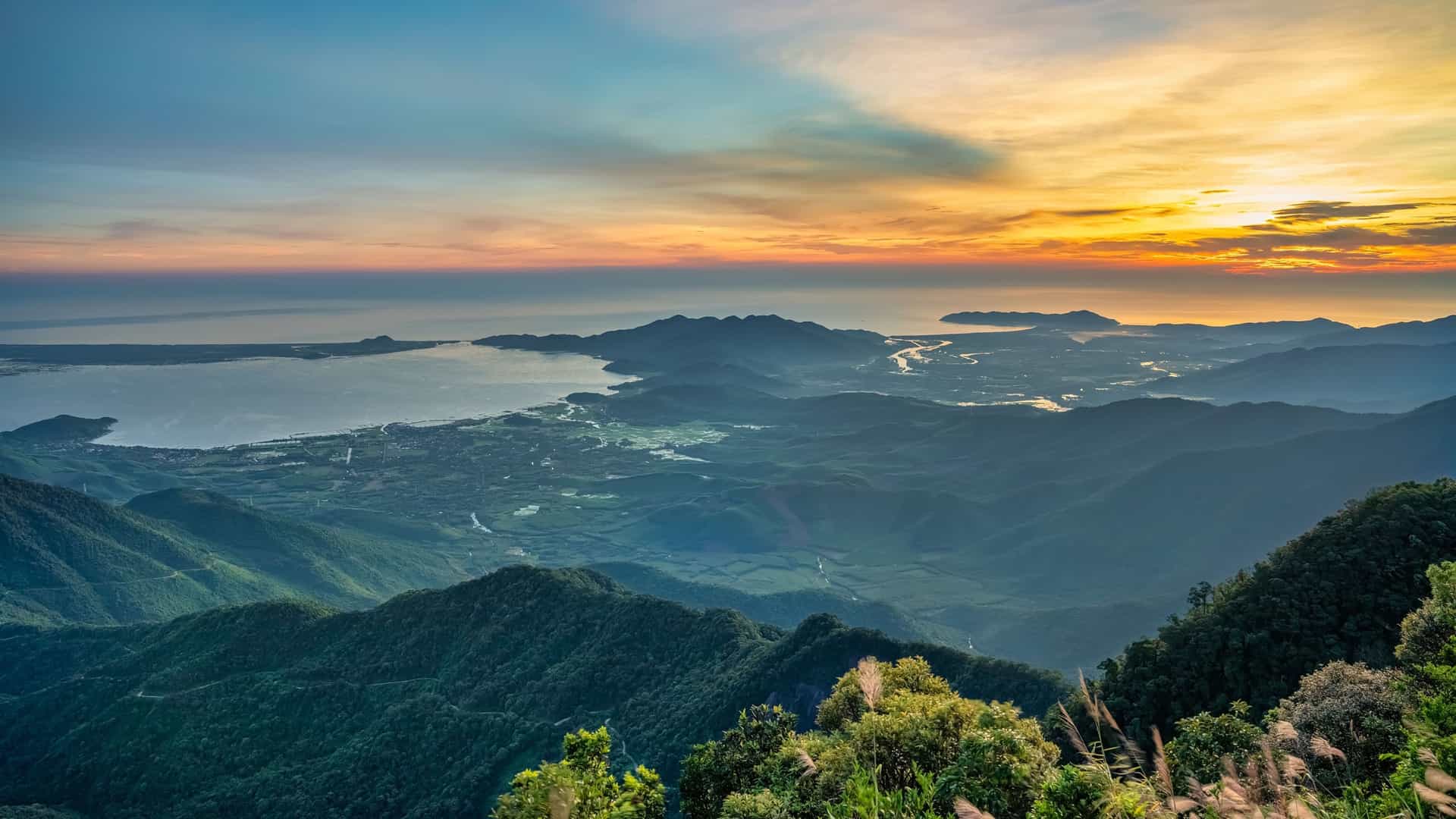 A dramatic aerial shot of the Hai Van Pass near Hue, Vietnam, with a bay and coastline visible between lush green mountains under a vibrant sunrise sky.